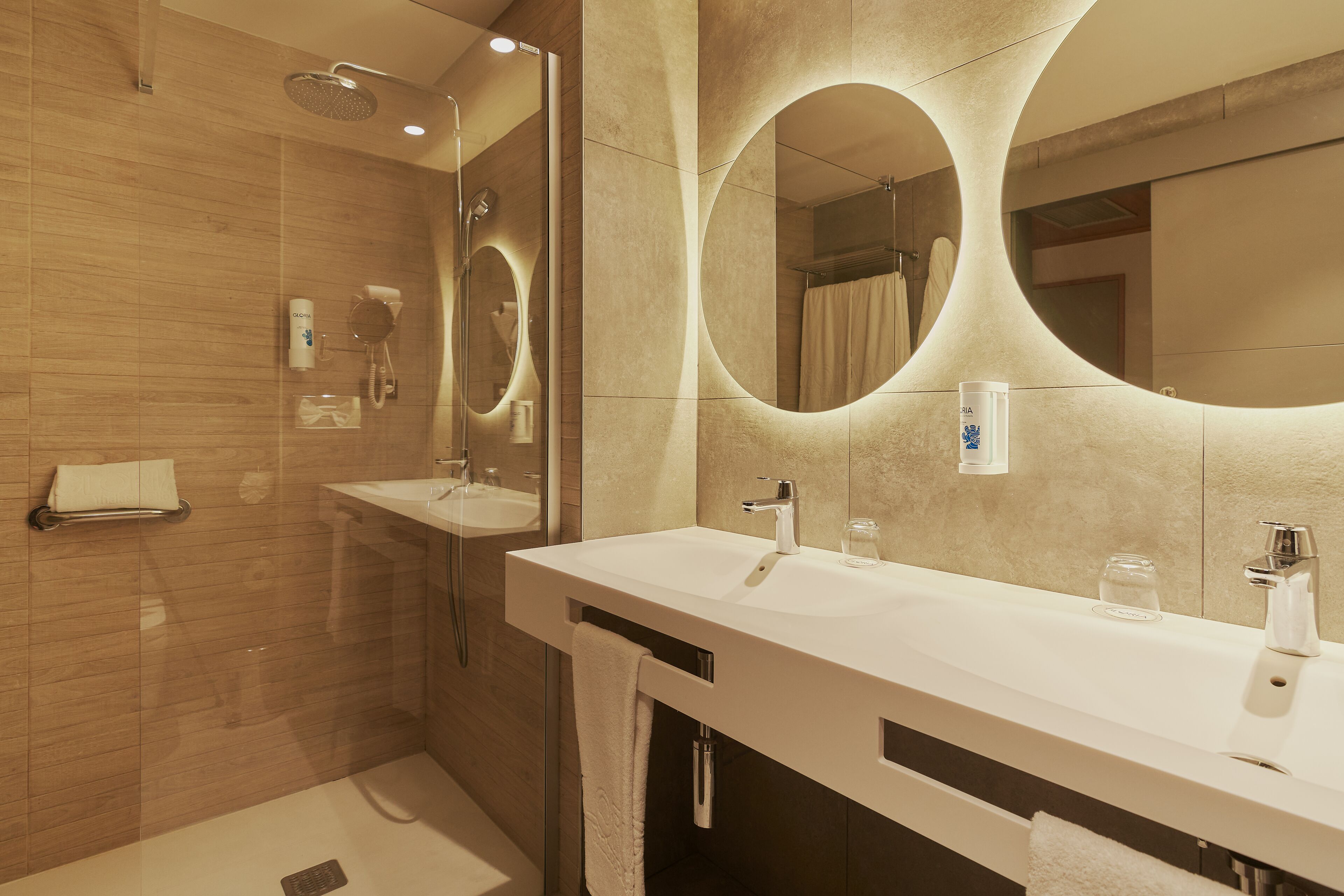 Une salle de bain moderne avec deux lavabos, une douche et un grand miroir doté d’un éclairage circulaire.