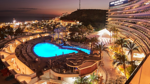 A hotel with a large swimming pool, multiple balconies, and a view of the surrounding cityscape.