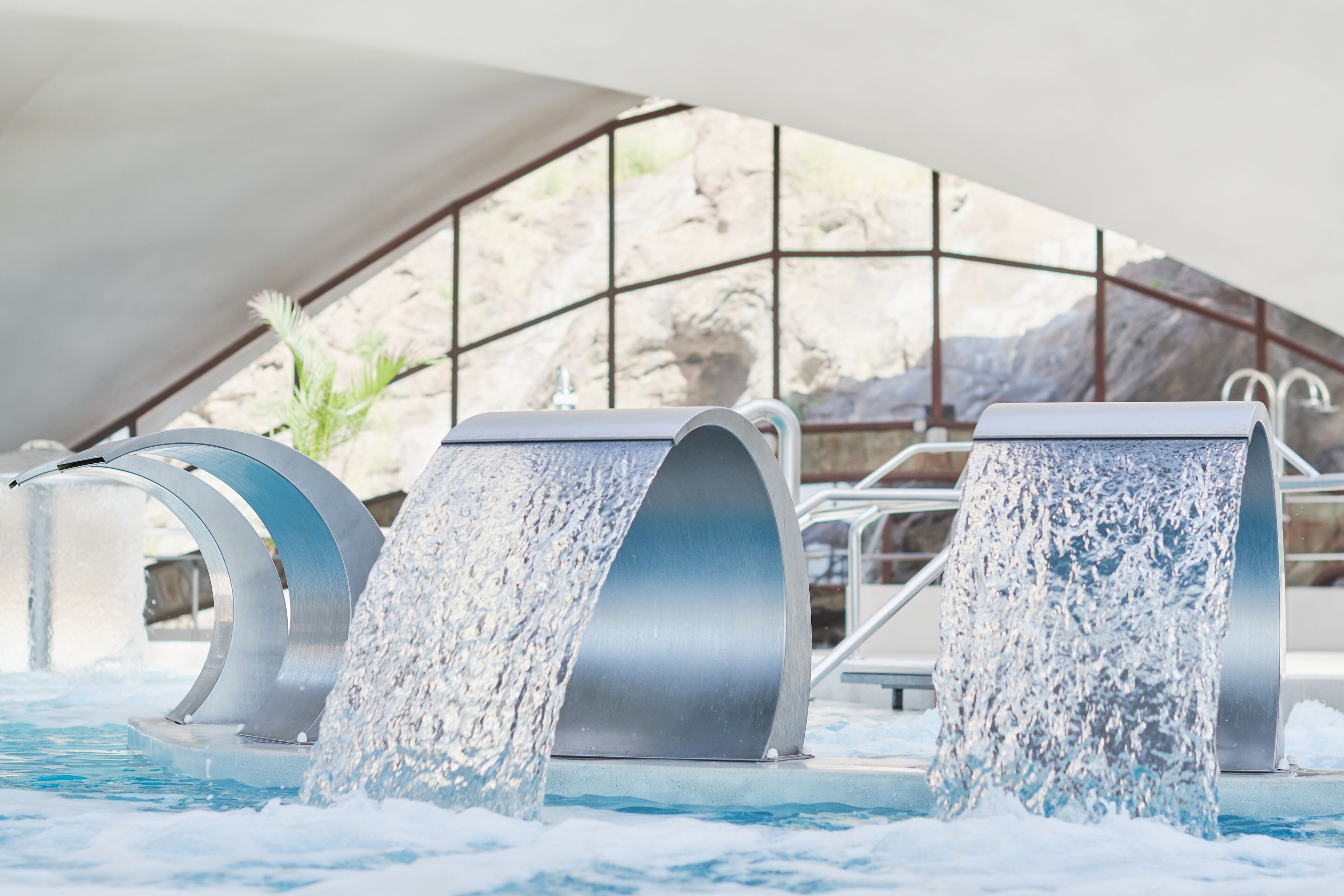 A pool with three water features and a view of a rocky landscape through a glass structure.