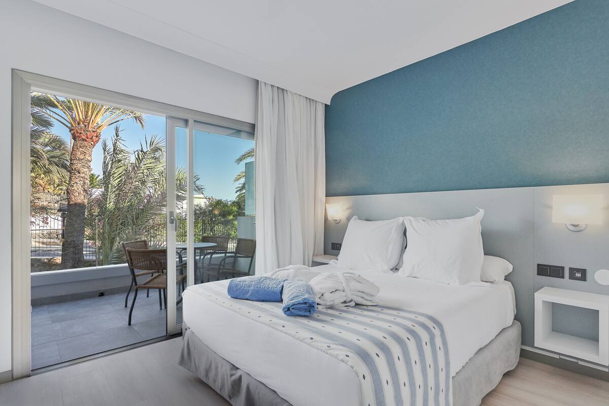 Une chambre d’hôtel moderne avec un grand lit, une table de chevet, une fenêtre donnant sur des palmiers et un balcon avec une chaise.