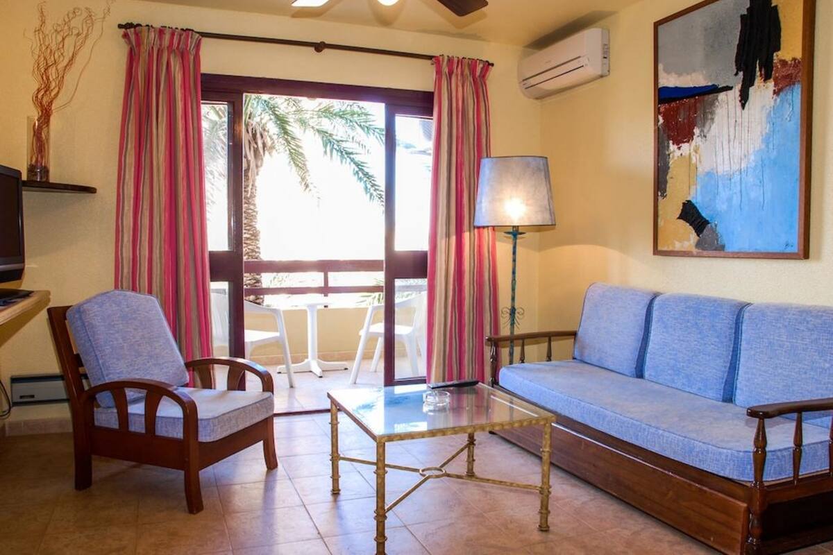 A living room with a sofa, a wooden chair, a glass coffee table, and a lamp. There is a window with curtains and an air conditioning unit on the wall.
