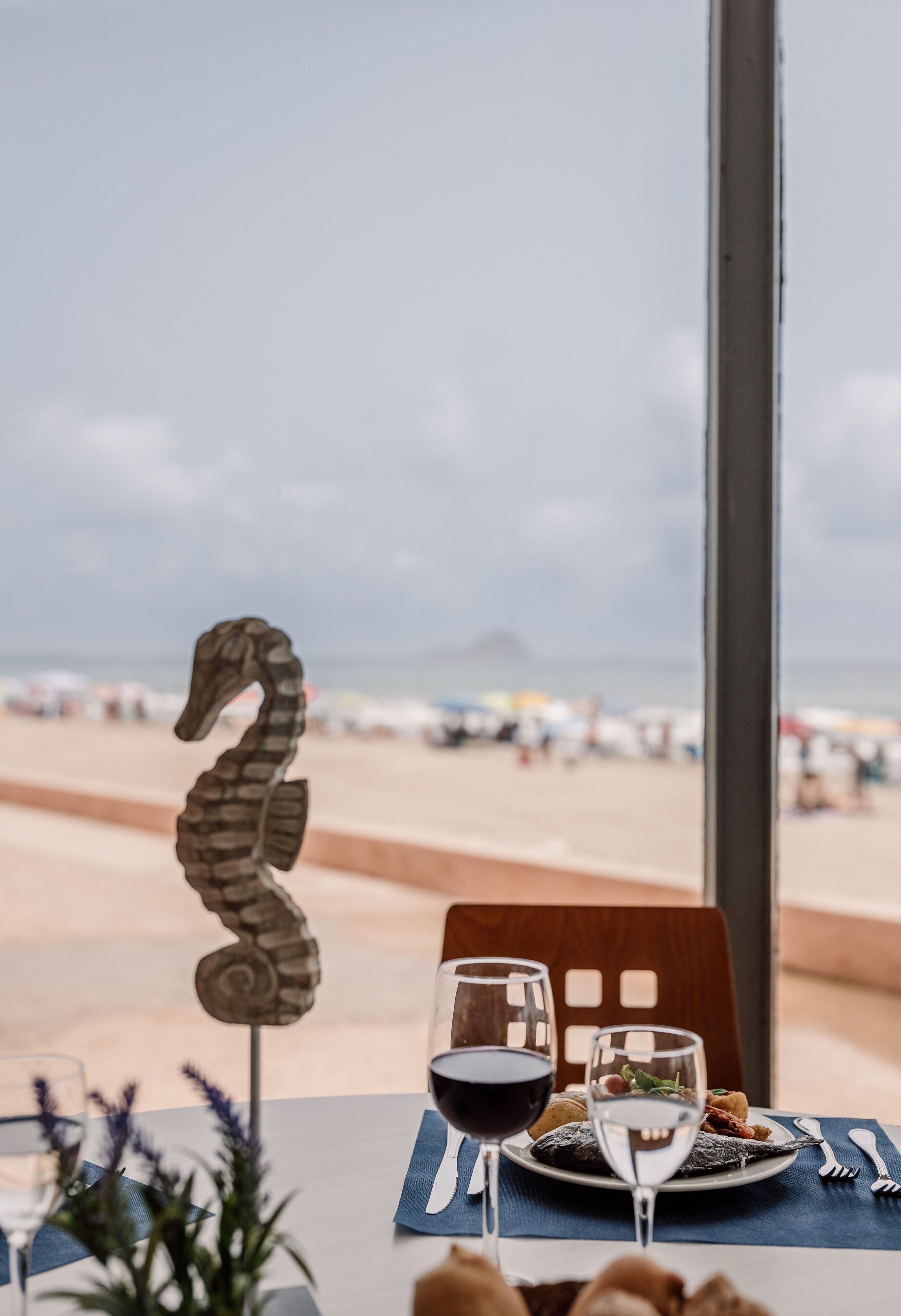 A table with a seahorse decoration, two glasses of red wine, and a plate of food with a view of the beach and ocean in the background.