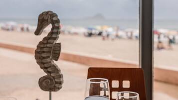 A table with a seahorse decoration, two glasses of red wine, and a plate of food with a view of the beach and ocean in the background.