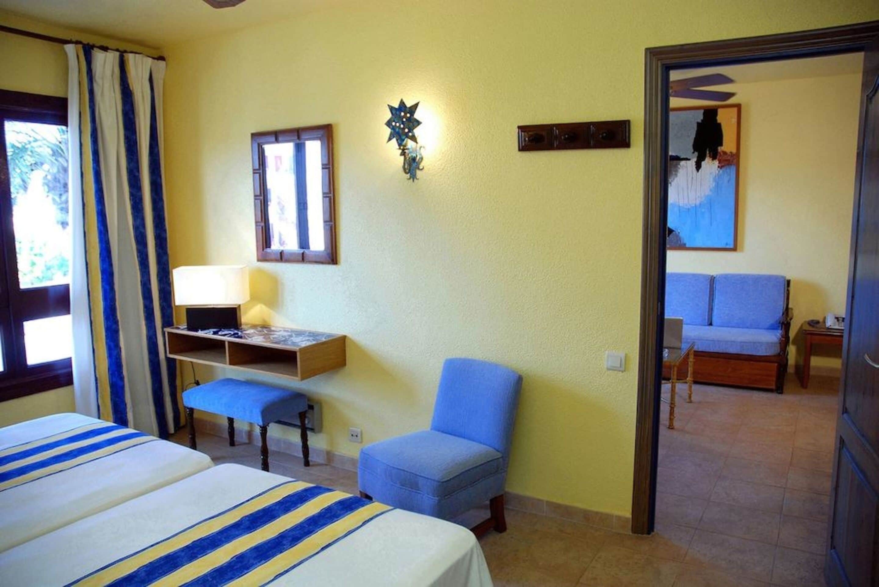 A hotel room with two beds, a desk with a lamp, a blue chair, and a bed with a blue cushion.