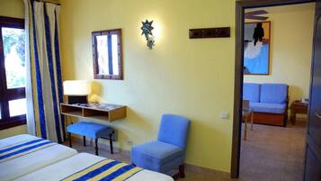 A hotel room with two beds, a desk with a lamp, a blue chair, and a bed with a blue cushion.