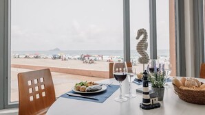 A table with a plate of food, two wine glasses, a lighthouse model, and a basket of bread, overlooking a beach with umbrellas and people. - Aparthotel Londres (San Javier)