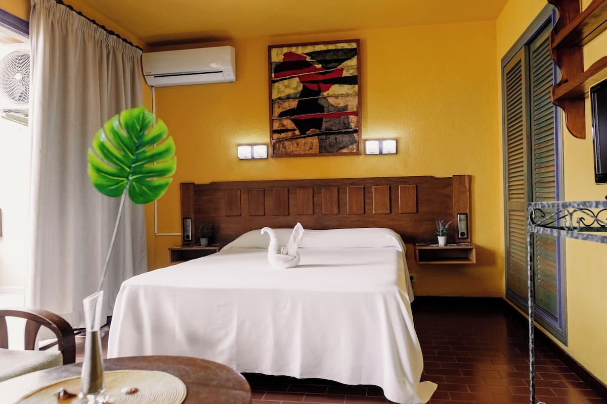 A hotel room with a bed, a wall-mounted air conditioner, a decorative painting, bedside lamps, and a potted plant.