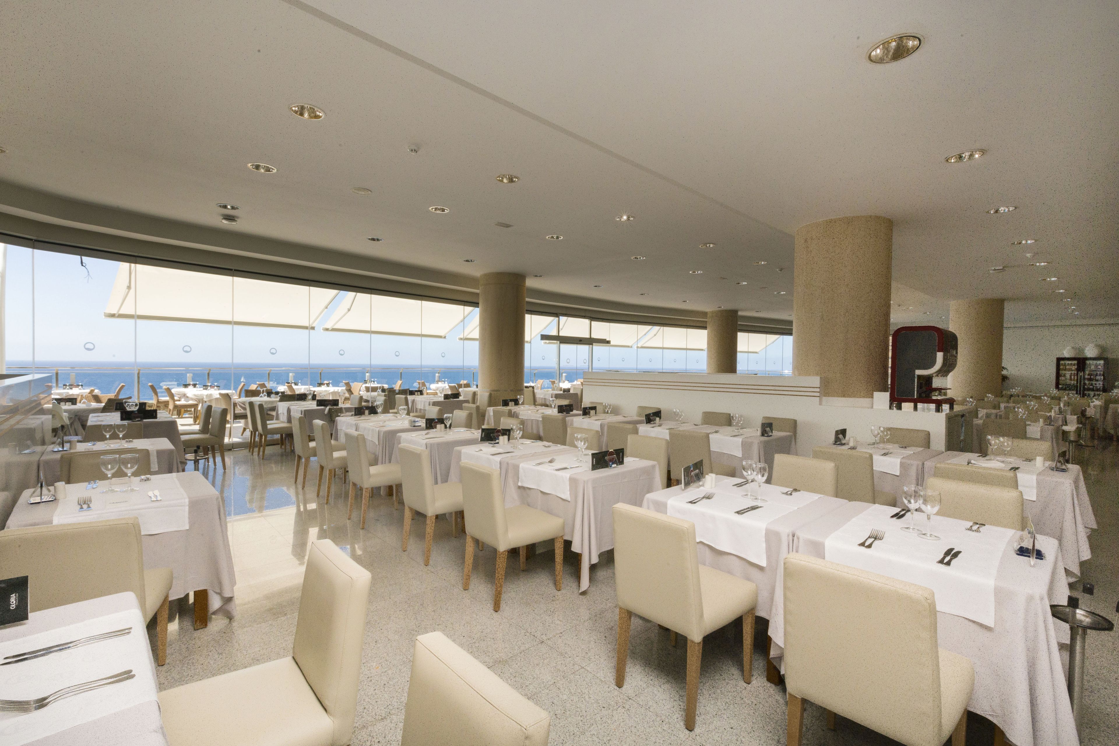 A spacious dining area with tables set for multiple guests, large windows offering a view of the sea, and a high ceiling with recessed lighting.