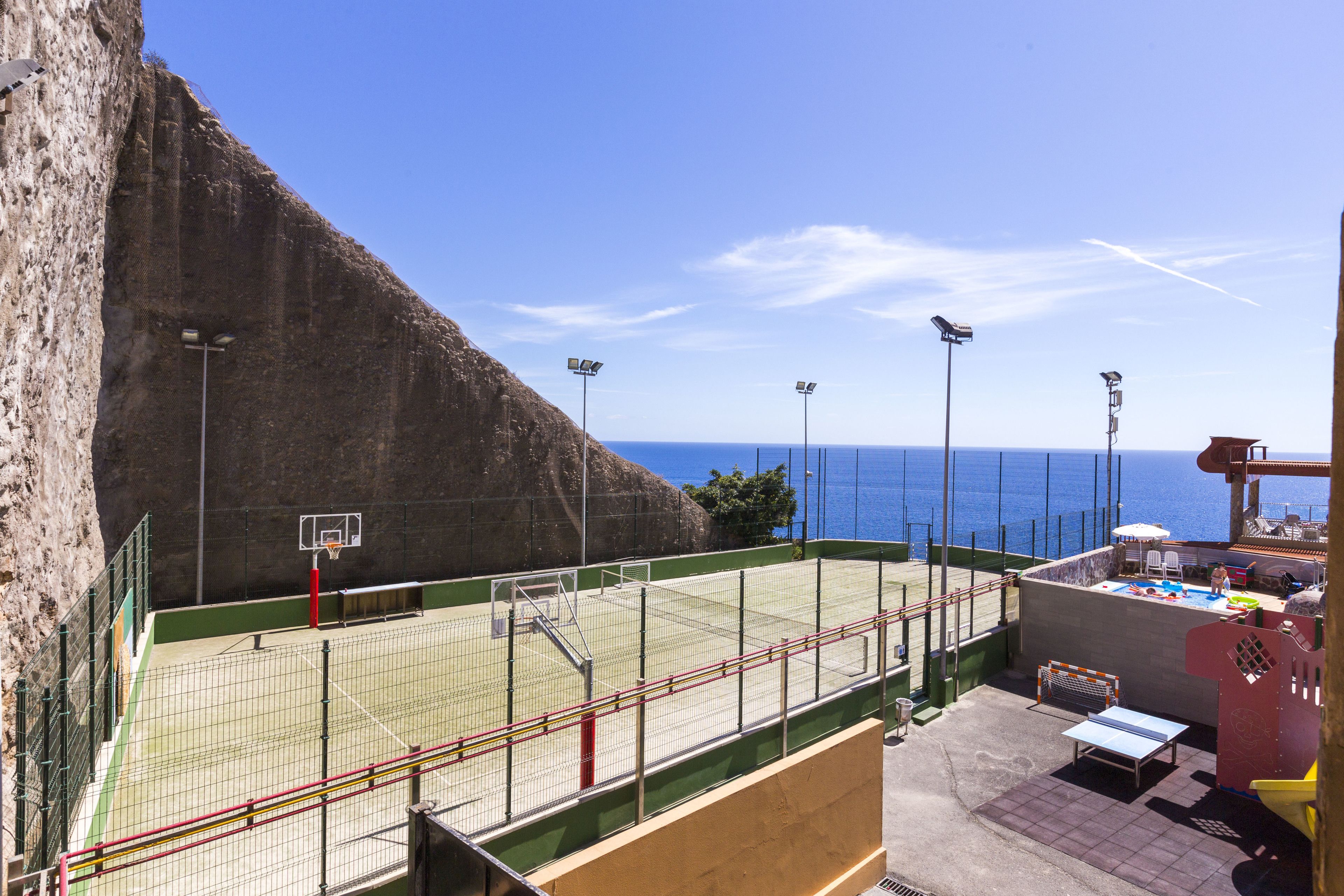 Una cancha deportiva al aire libre con un aro de baloncesto, junto a un acantilado y el mar.