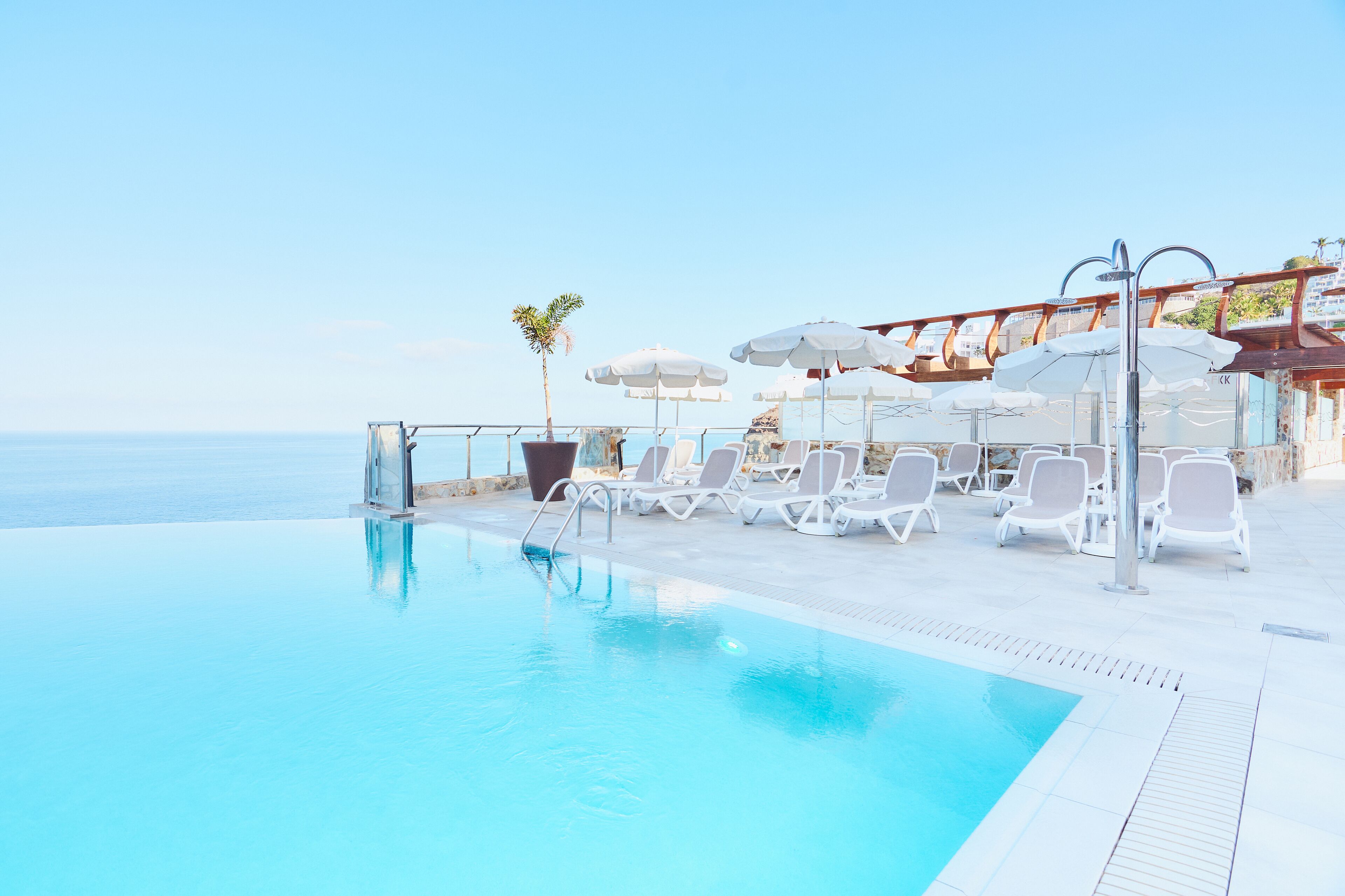 Un espace piscine avec des chaises longues blanches et des parasols, un palmier et une vue sur la mer.