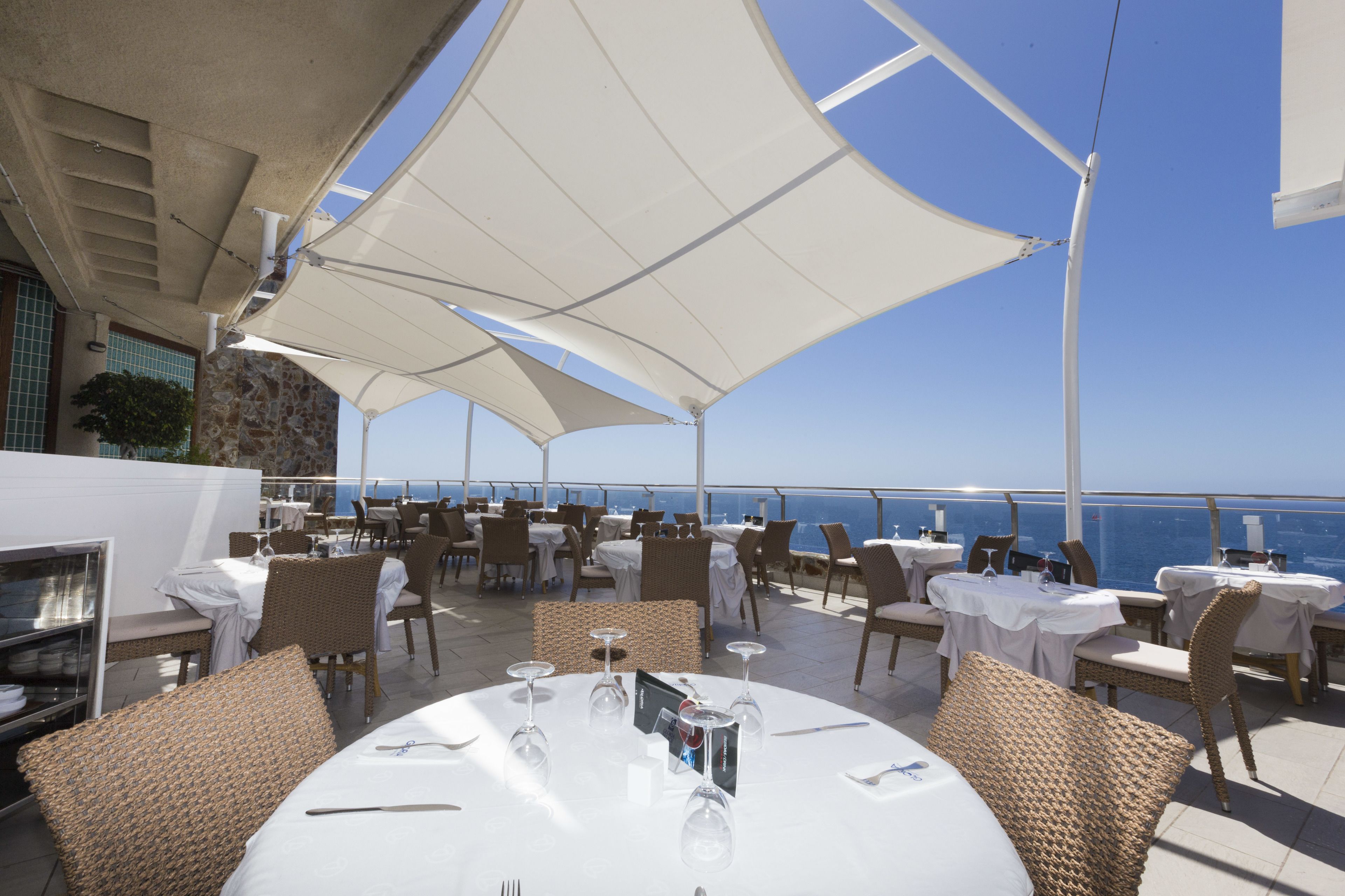 An outdoor dining area with tables and chairs, large white umbrellas, and a view of the sea.