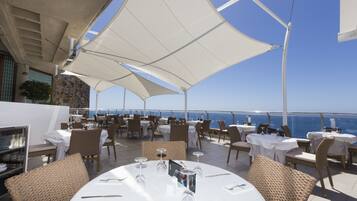 An outdoor dining area with tables and chairs, large white umbrellas, and a view of the sea.