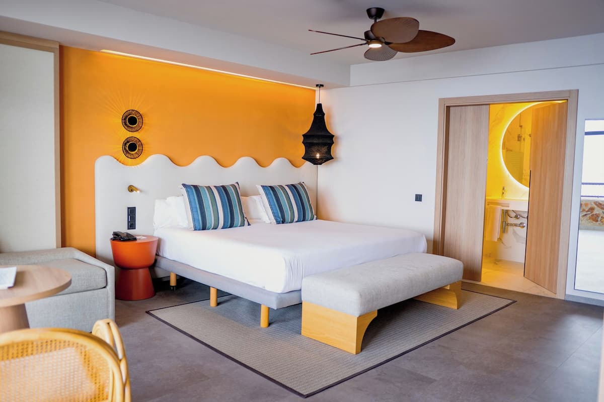 Ein modernes Hotelzimmer mit einem Bett, einem kleinen Tisch, einem Sessel, einem Deckenventilator und einer Tür zu einem Badezimmer.
