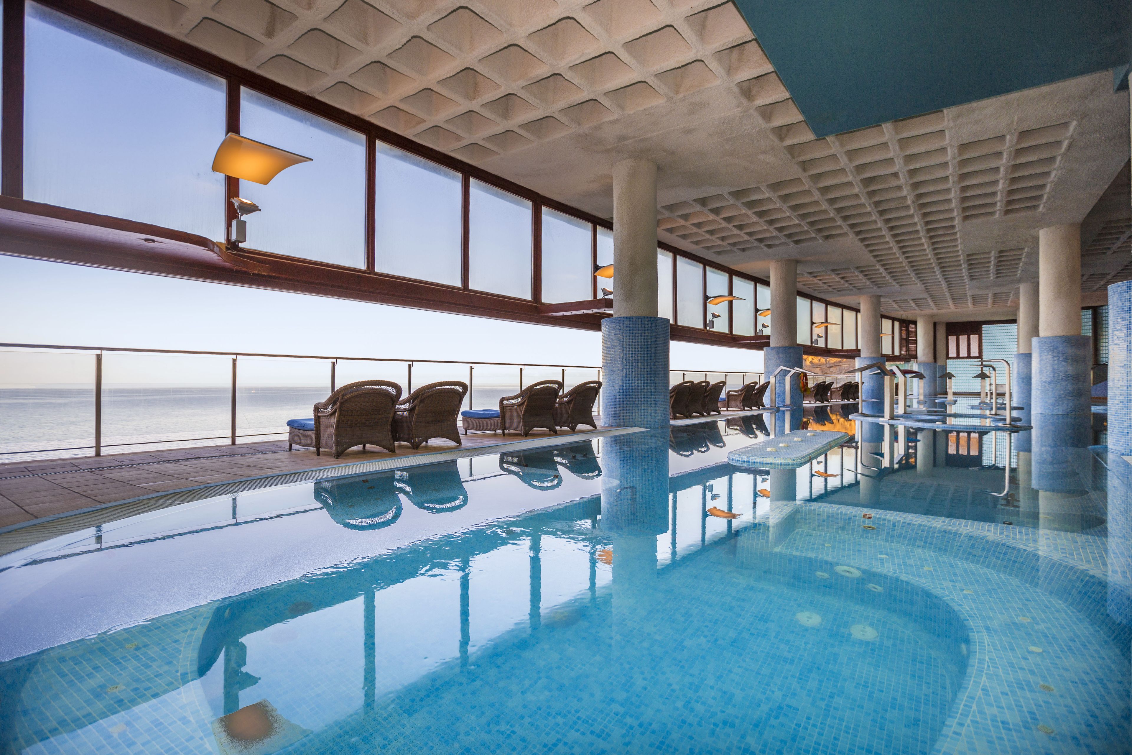An indoor swimming pool with clear views of the ocean, lounge chairs, and a tiled ceiling.
