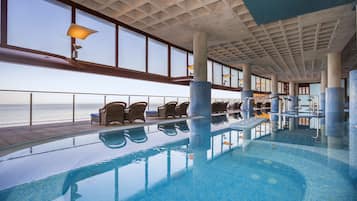 An indoor swimming pool with clear views of the ocean, lounge chairs, and a tiled ceiling.