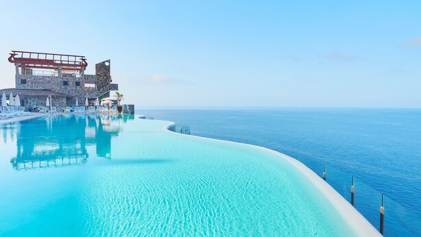 Una piscina infinita con agua azul cristalina, una orilla de arena blanca y un edificio con un diseño arquitectónico singular al fondo.