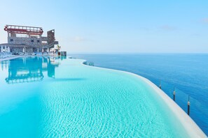 A infinity pool with clear blue water, a white sandy edge, and a building with a unique architectural design in the background. - Gloria Palace Amadores Thalasso & Hotel (Mogan)