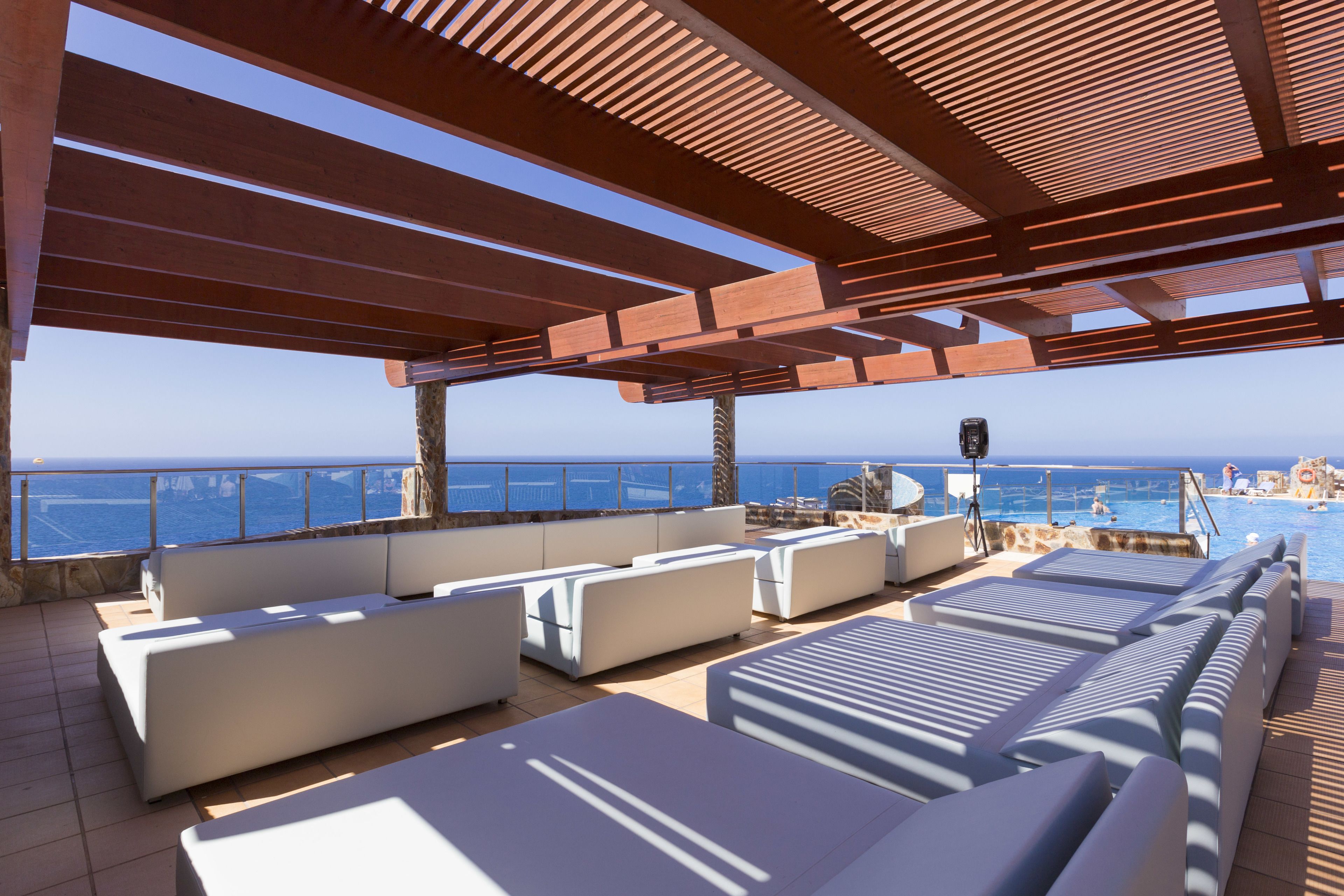 Terraza con sofás blancos y sillones de sol, con vistas a una piscina y al mar.