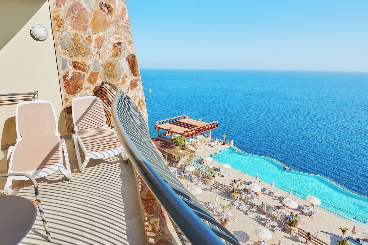A balcony with lounge chairs overlooking a pool and the sea.
