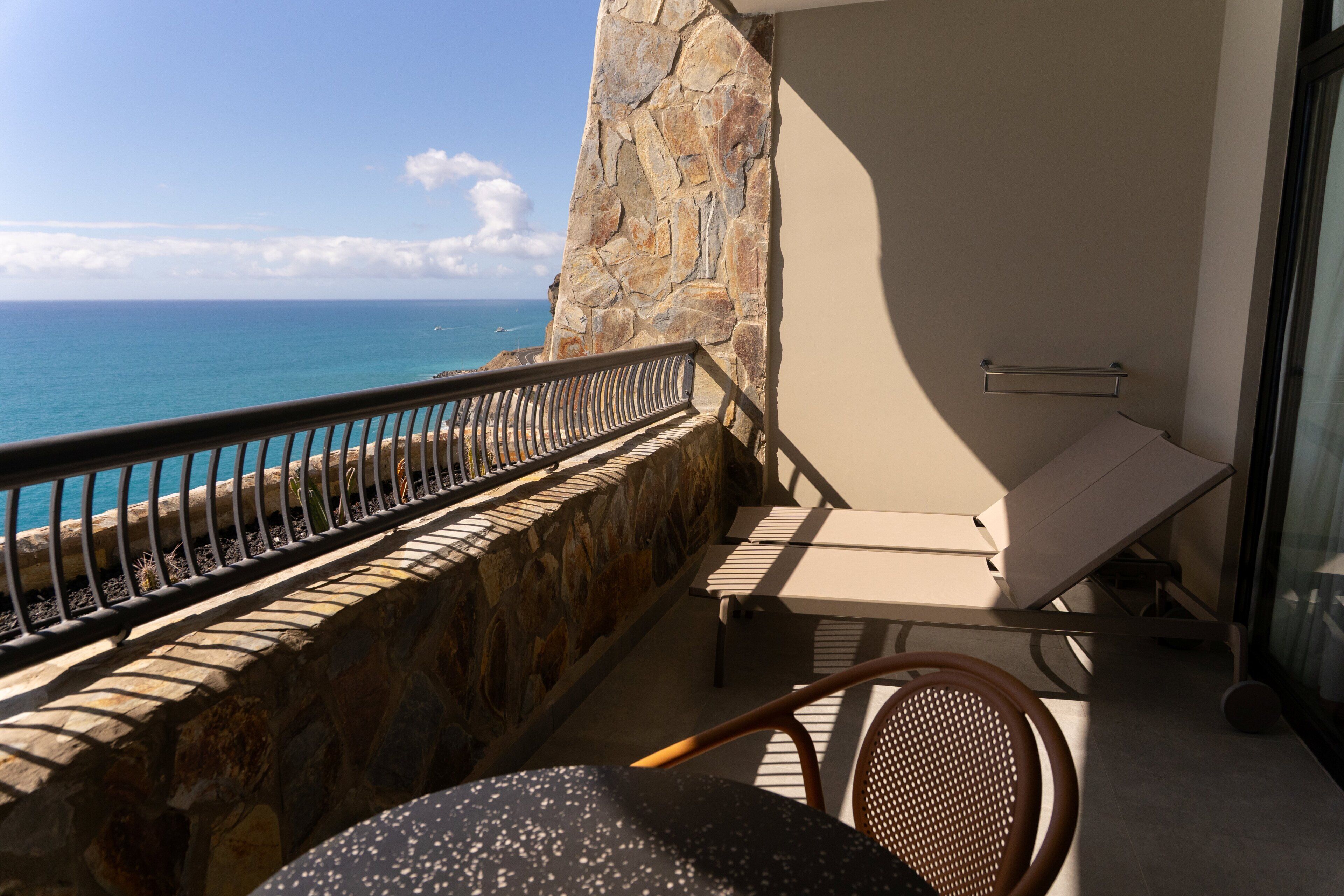A balcony with a stone wall, a metal railing, a lounge chair, and a table.