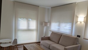 A modern living room with a beige sofa, a wooden coffee table, and a small white table. There are two white chairs, a floor lamp, and large windows with blinds. - Hotel Arias Aeropuerto (Castrillon)
