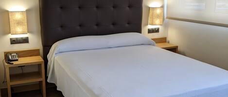 Una cama bien hecha con un cabecero oscuro, dos mesitas de noche con lámparas y suelo de madera.