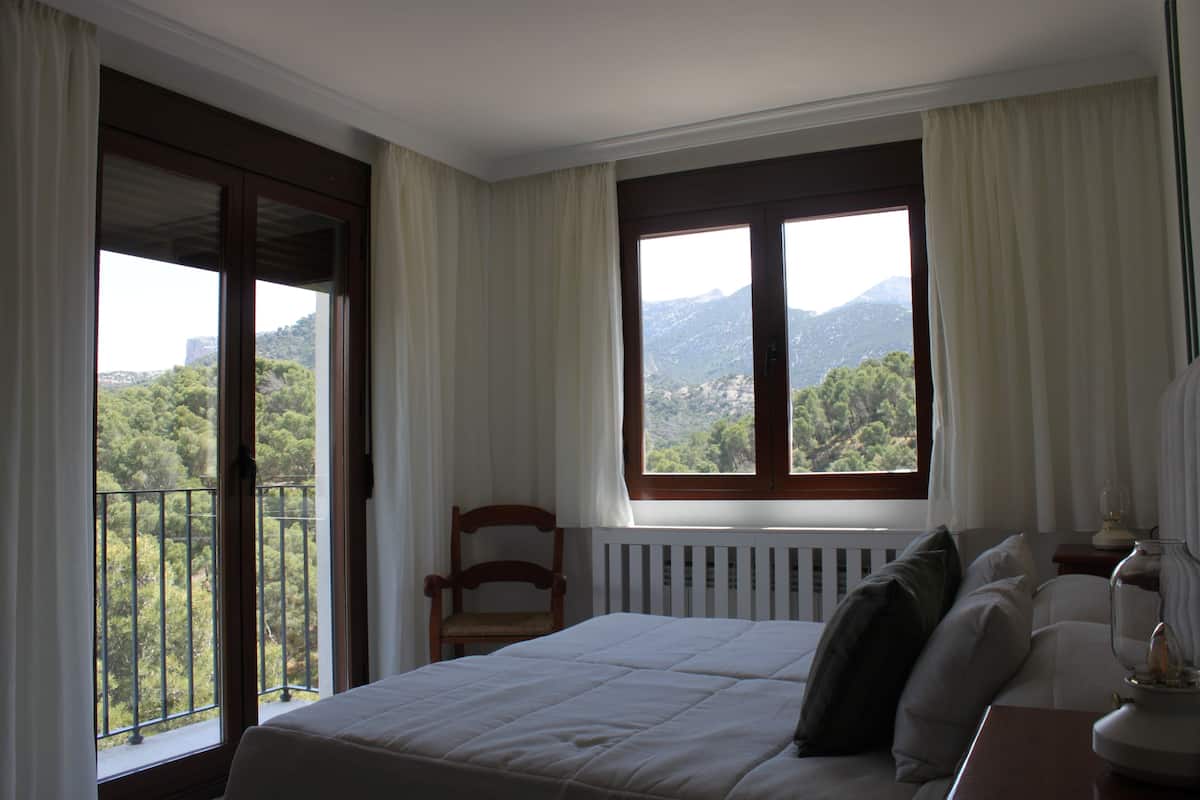 A bedroom with a bed, a chair, a nightstand, and a view of greenery and mountains through the window.