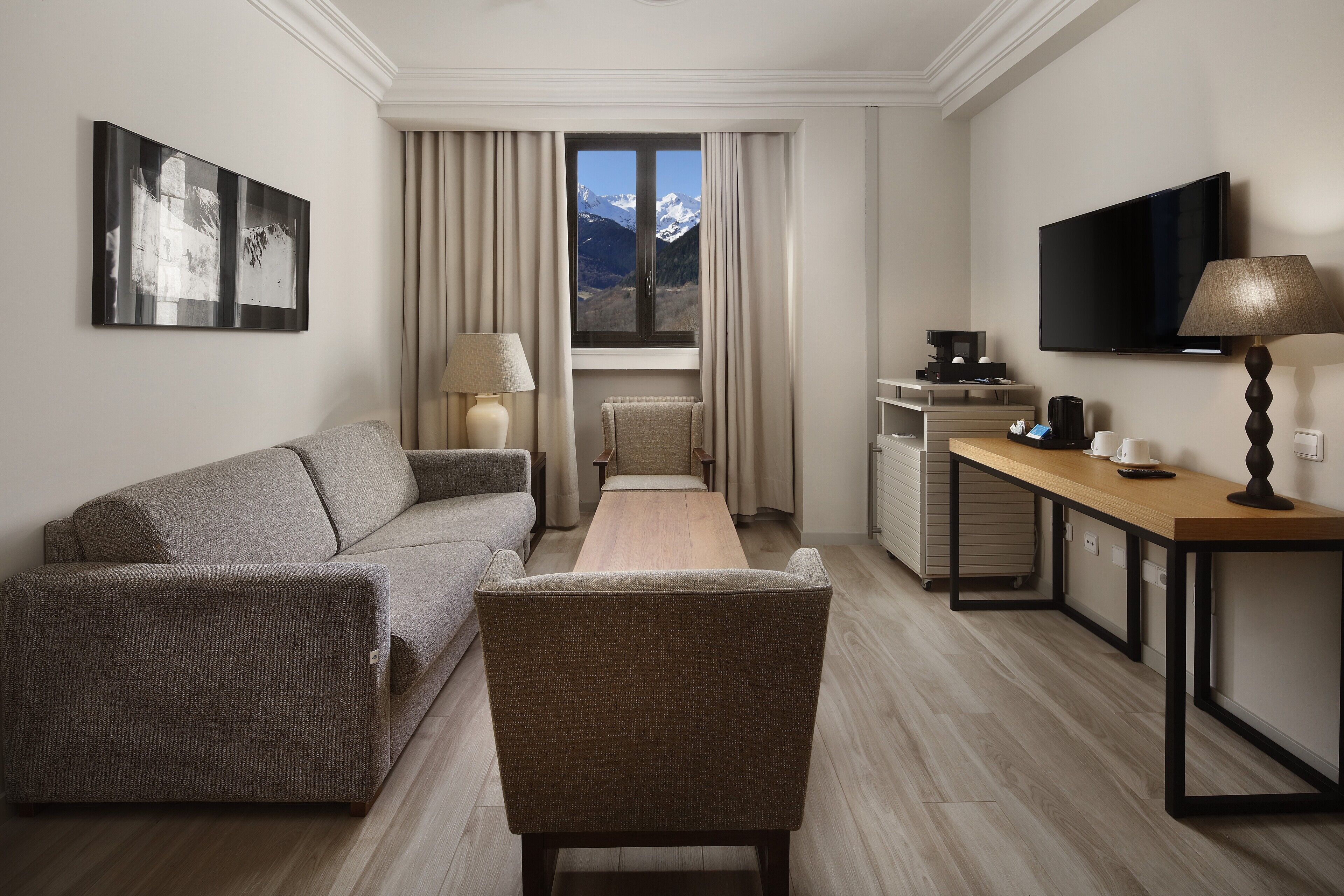 A modern hotel room with a sofa, a chair, a desk, a TV, a lamp, and a window with a mountain view.