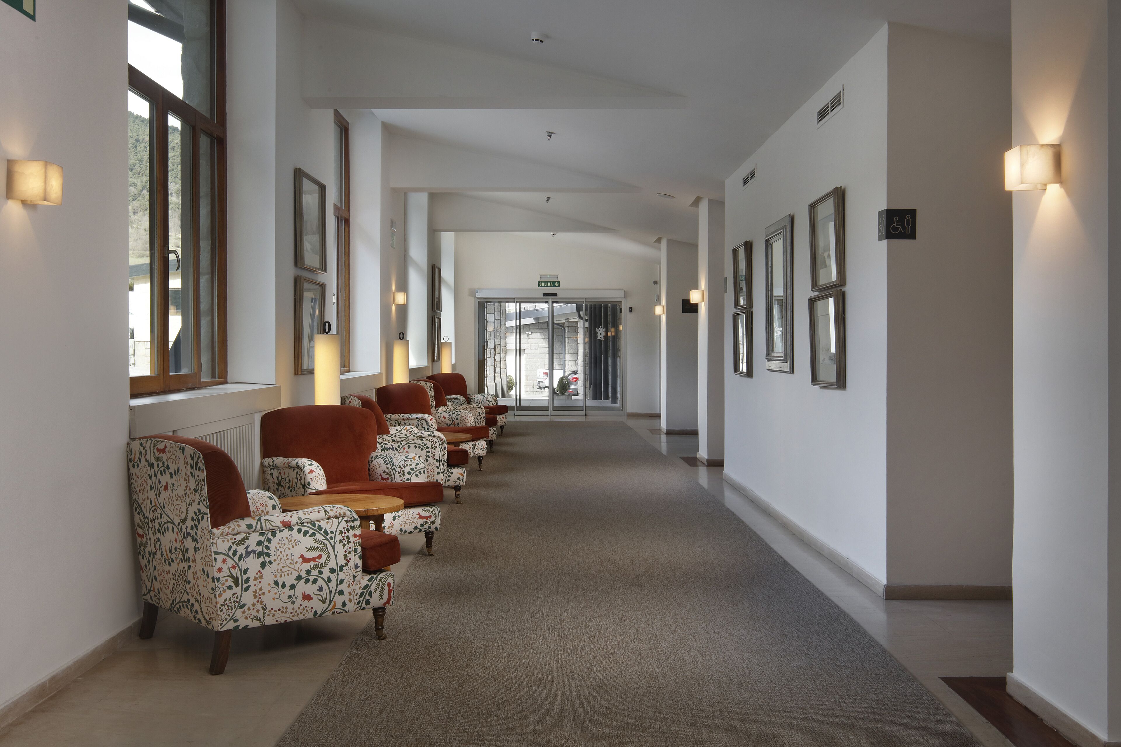 A hotel corridor with patterned armchairs, framed pictures on the walls, and a carpeted floor.
