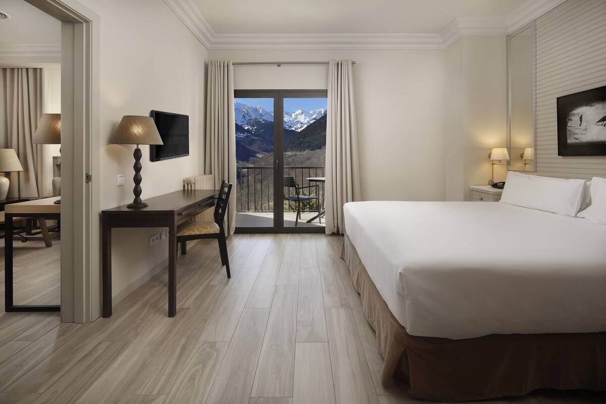 A hotel room with a bed, a desk, a chair, a table, a lamp, a painting, a window with a mountain view, and a sliding door.