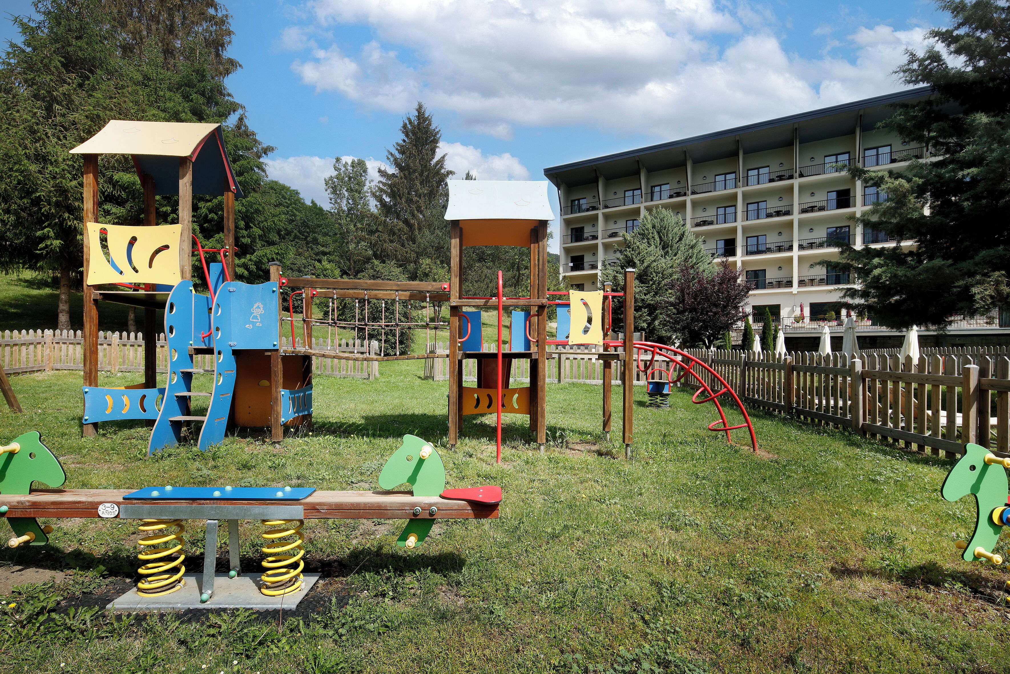 Un parque infantil con diversas estructuras de juego, una cerca de madera y un edificio con múltiples balcones al fondo.