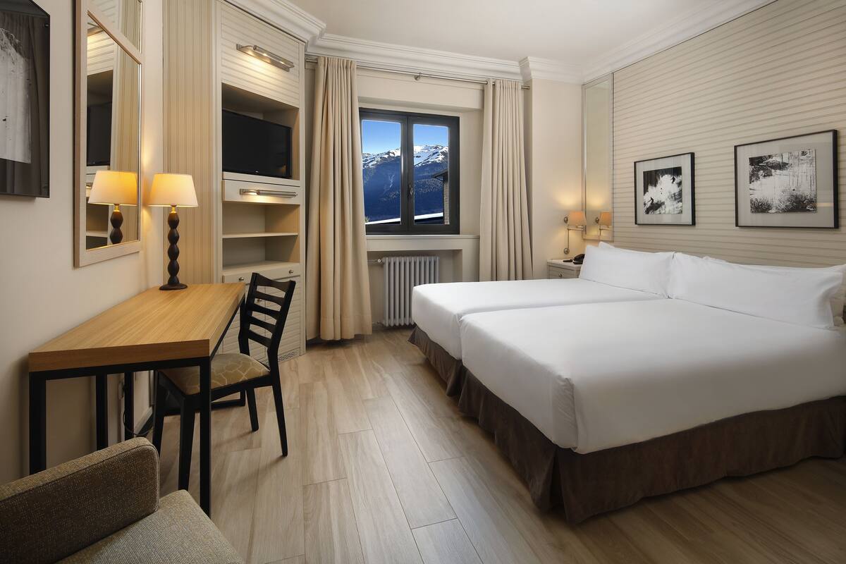A hotel room with a bed, a desk, a chair, a TV, a window with a mountain view, and two framed pictures on the wall.