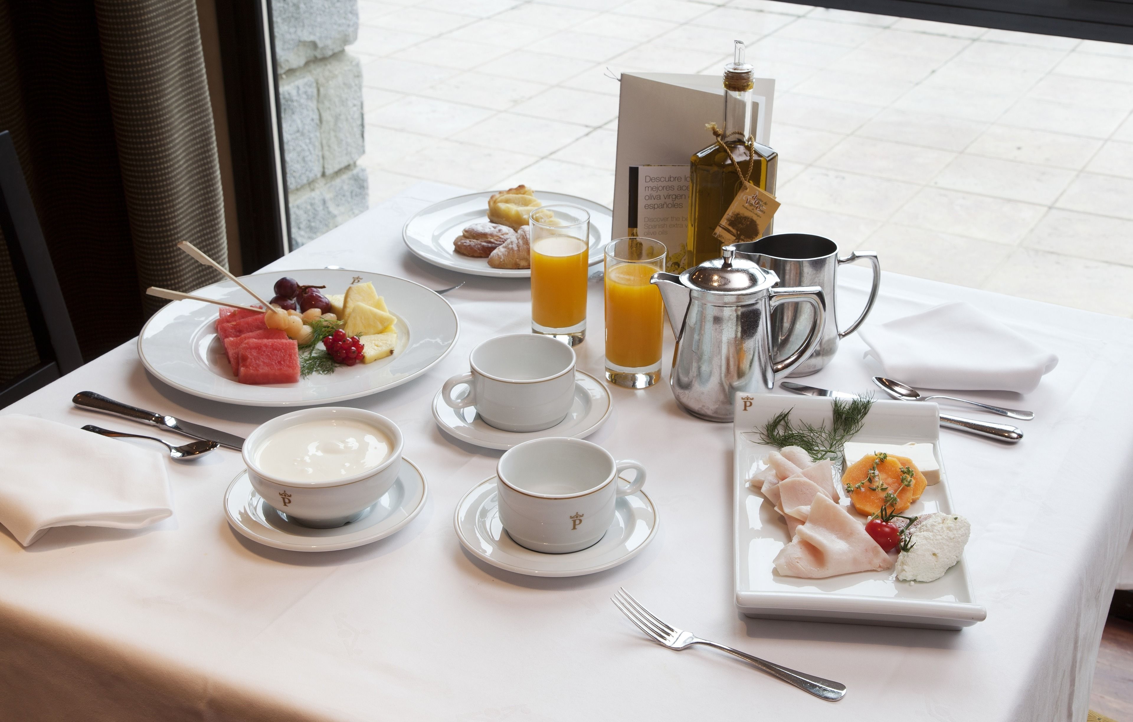 A breakfast table set with plates of fruit, yogurt, and pastries, along with cups and a coffee pot.