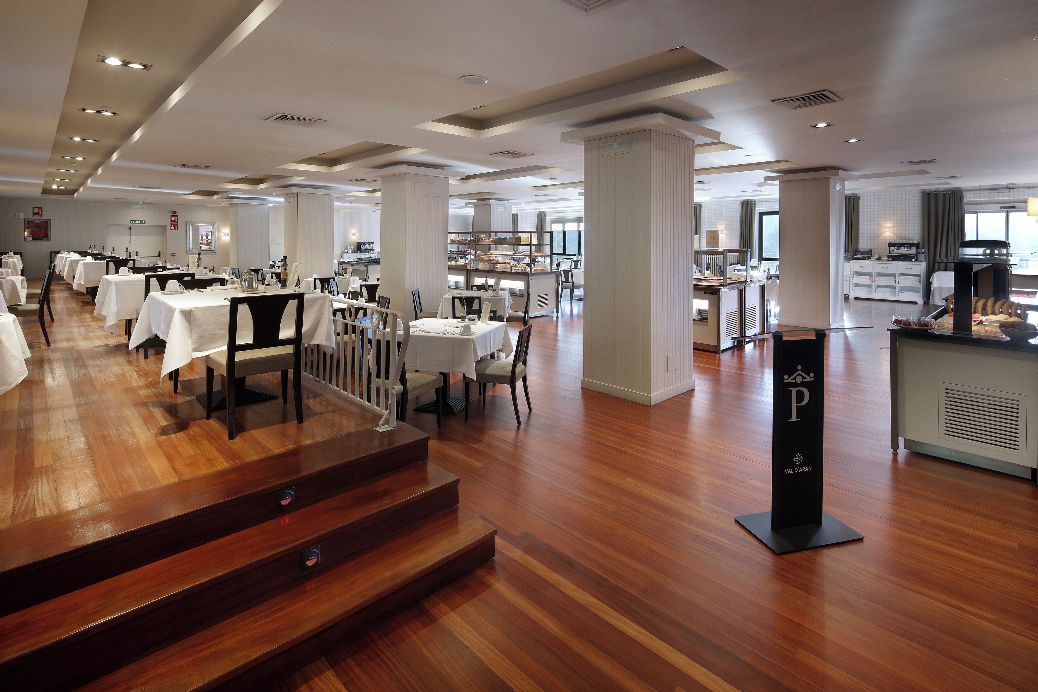 Amplio comedor de hotel con varias mesas y sillas, una escalera central y un mostrador tipo buffet.