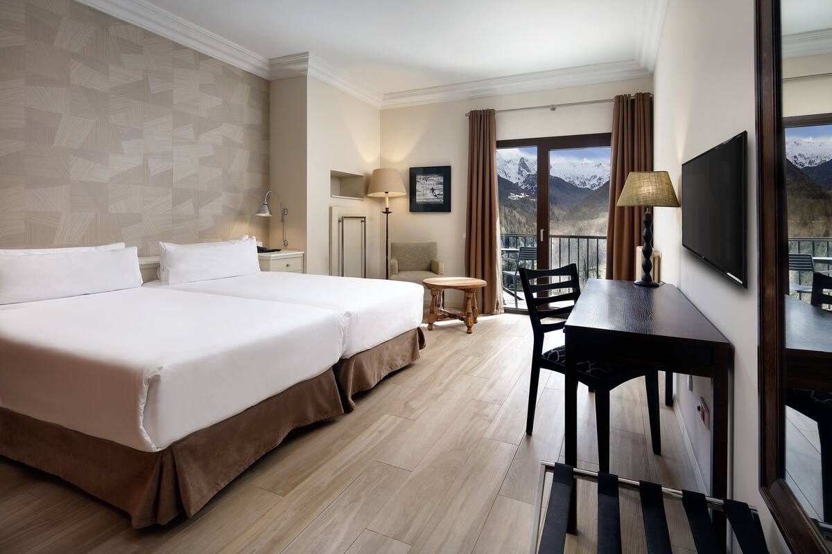 A hotel room with a large bed, a desk, a chair, a TV, and a balcony with a mountain view.