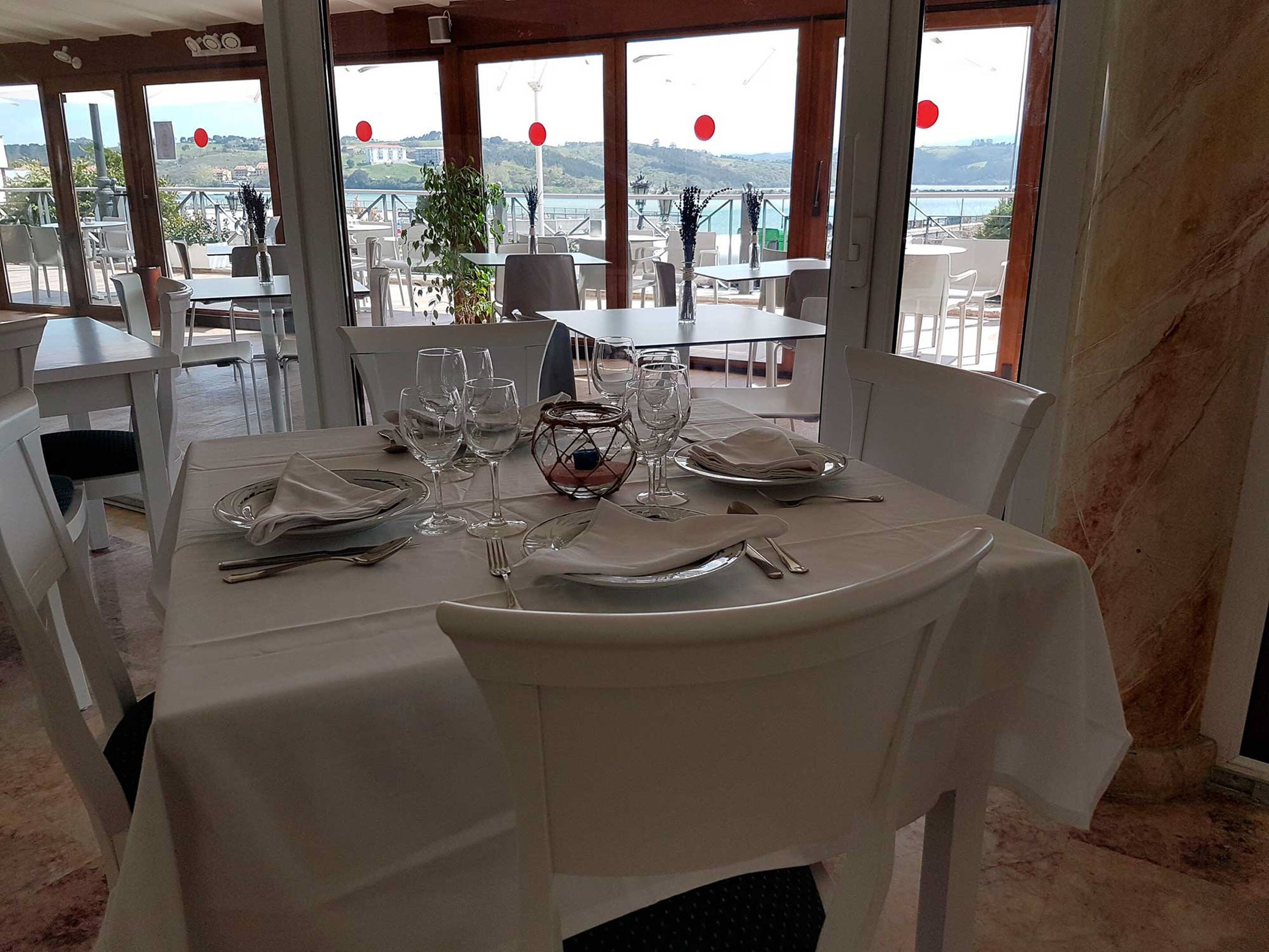 Mesa de comedor con mantel blanco, platos, cubiertos y copas de vino, con vista al mar y colinas al fondo.