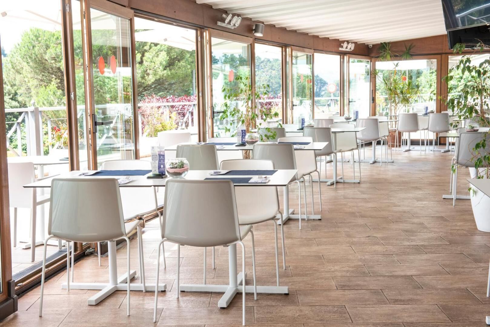 A spacious outdoor dining area with tables and chairs, large windows, and potted plants.