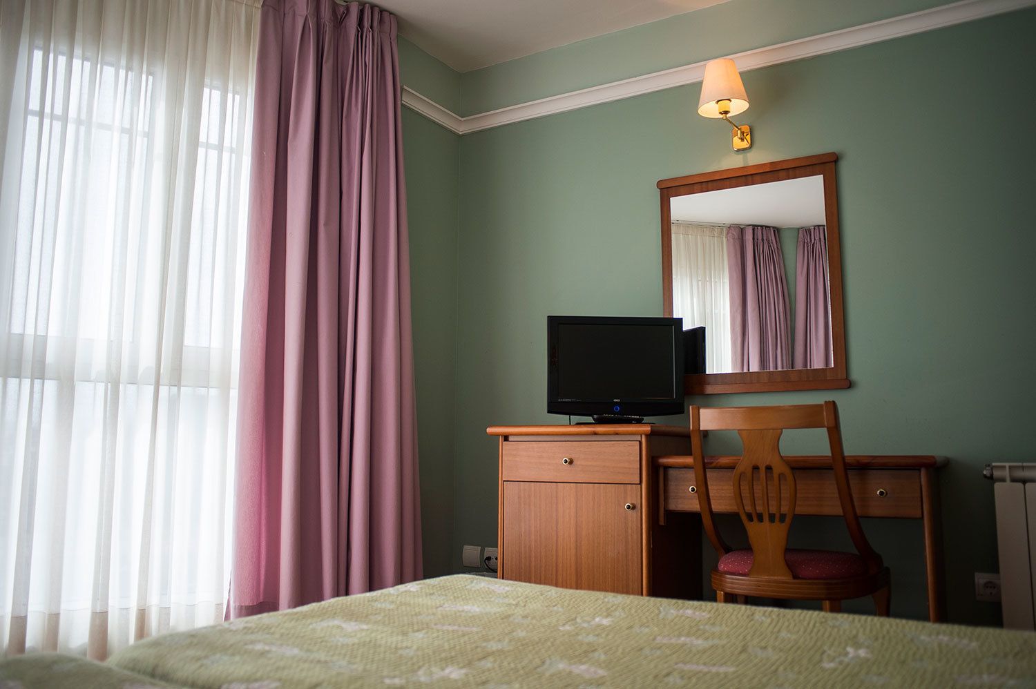 A hotel room with a bed, a wooden desk with a television, a chair, a mirror, and a window with curtains.