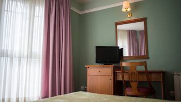 A hotel room with a bed, a wooden desk with a television, a chair, a mirror, and a window with curtains.