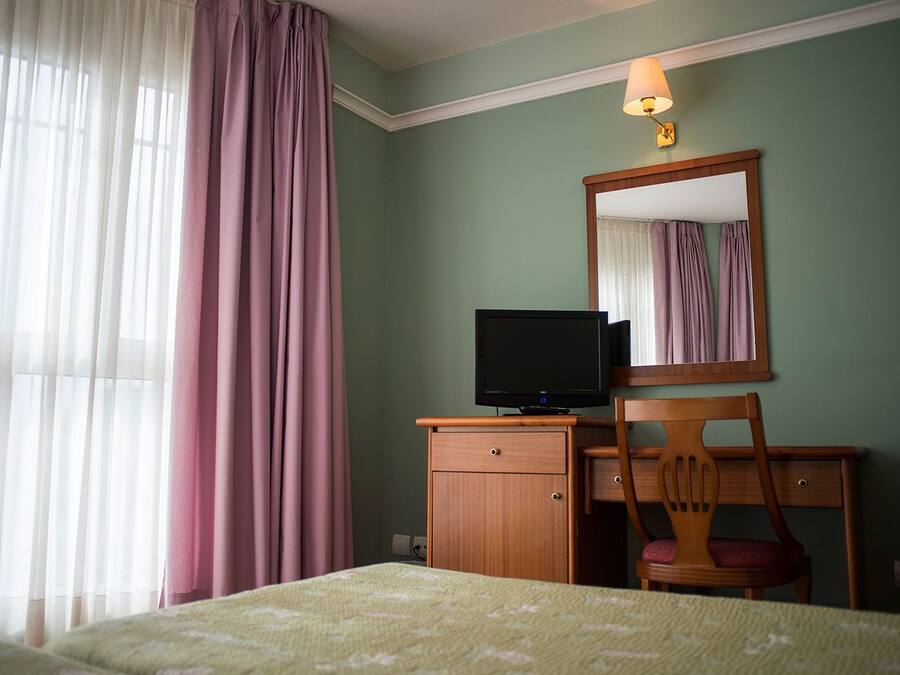 A hotel room with a bed, a wooden desk with a television, a chair, a mirror, and a window with curtains.