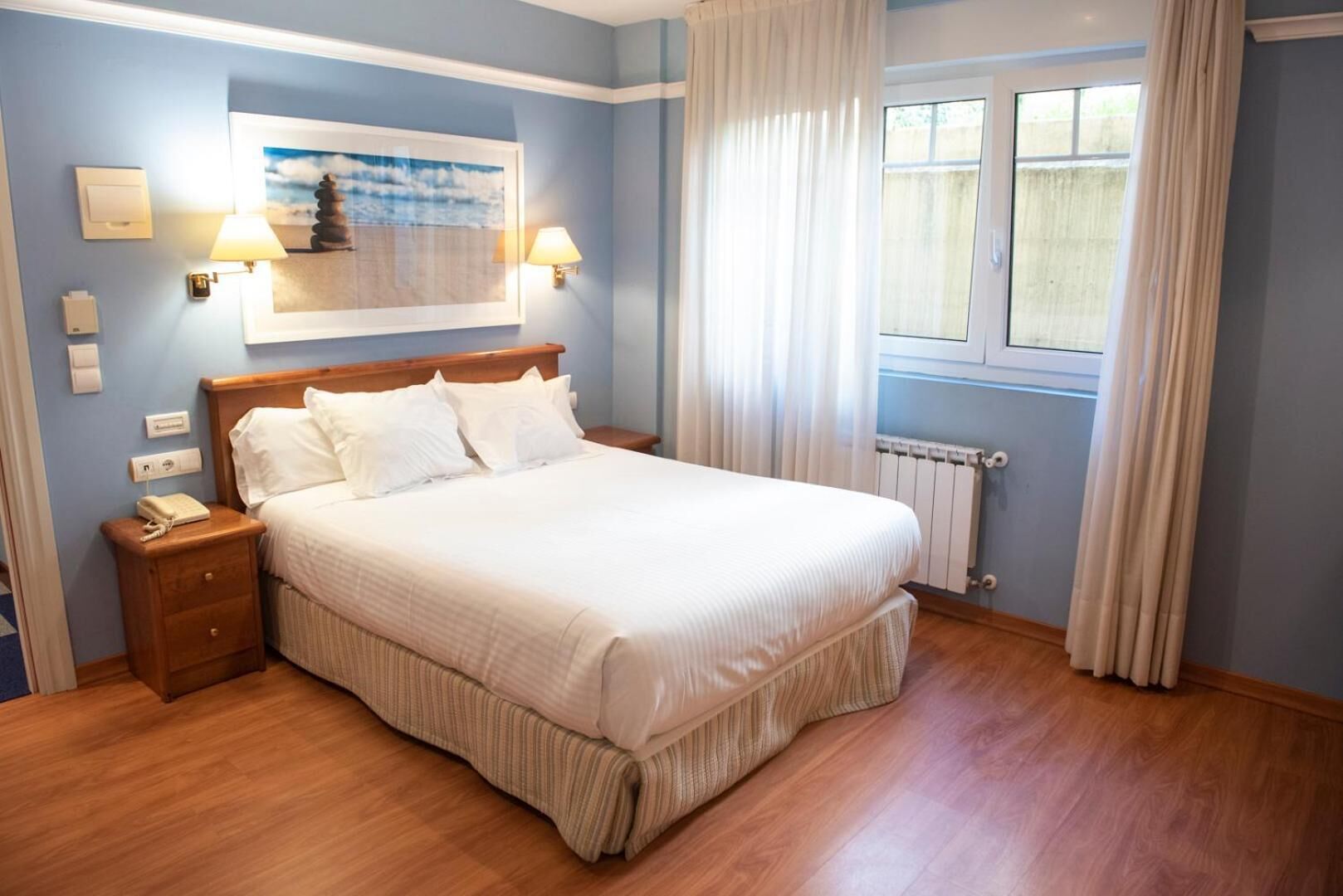 A neatly made bed in a room with a wooden headboard, a bedside table, a nightstand, a framed picture on the wall, a window with curtains, and a radiator.