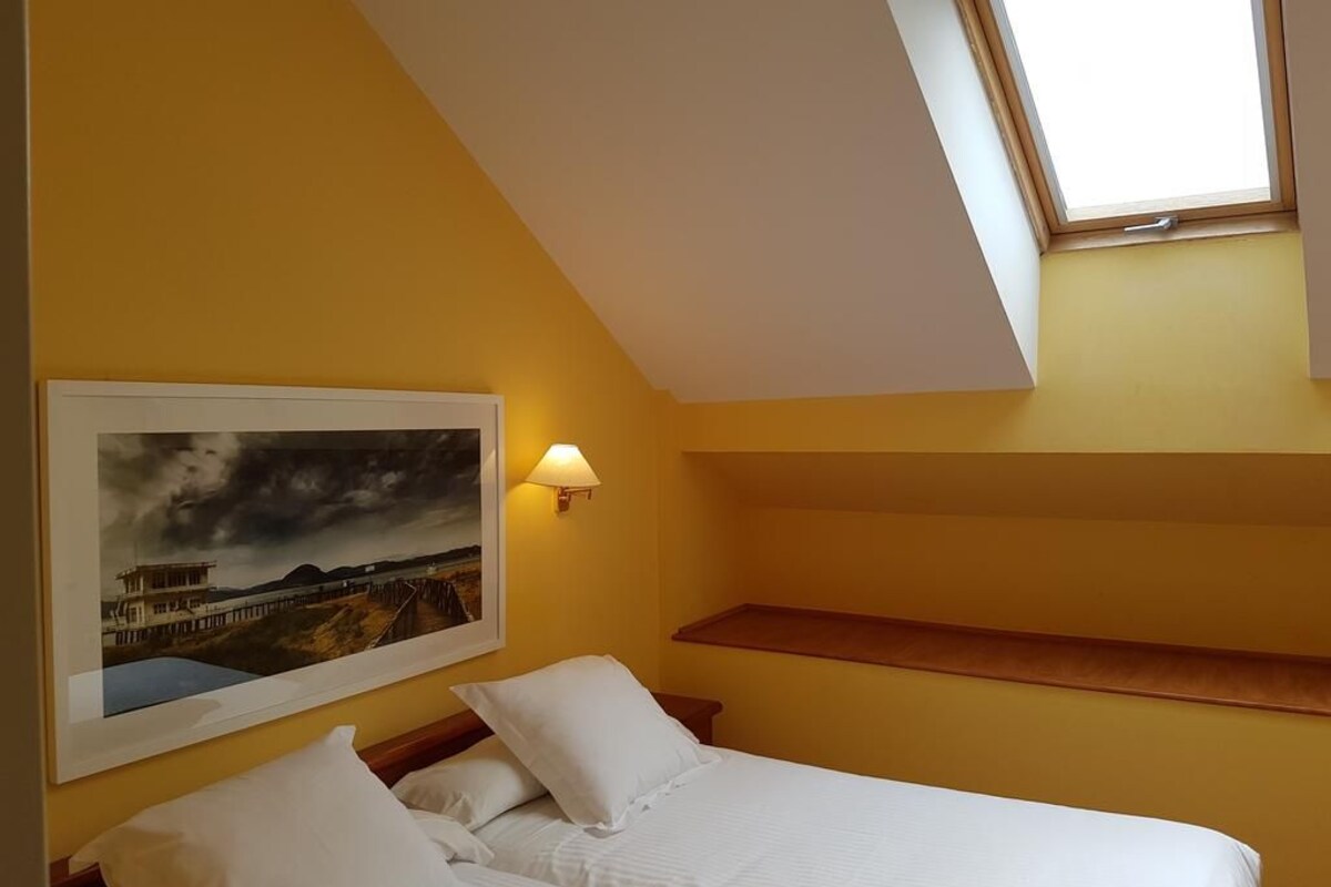 A bedroom with a bed, a painting, a nightstand, a lamp, and a skylight.