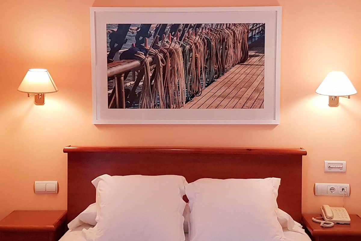 A hotel room with a bed, two bedside lamps, a framed picture of a ship's deck, and a telephone on the nightstand.
