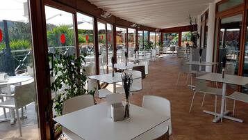 A covered outdoor dining area with white tables and chairs, large windows, and potted plants.