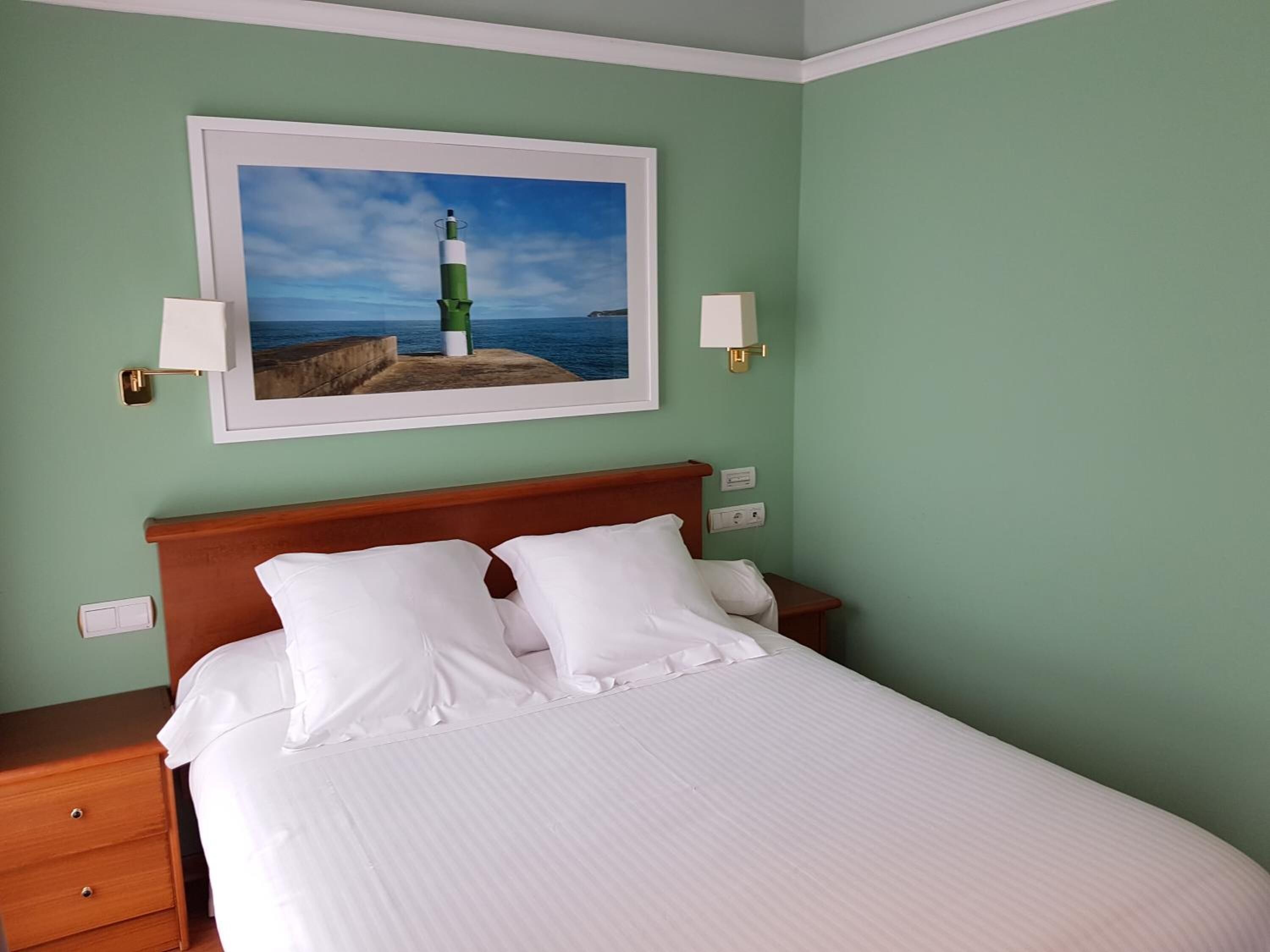 Una cama bien hecha con sábanas blancas en una habitación con paredes verdes, un cuadro de un faro y lámparas de noche.