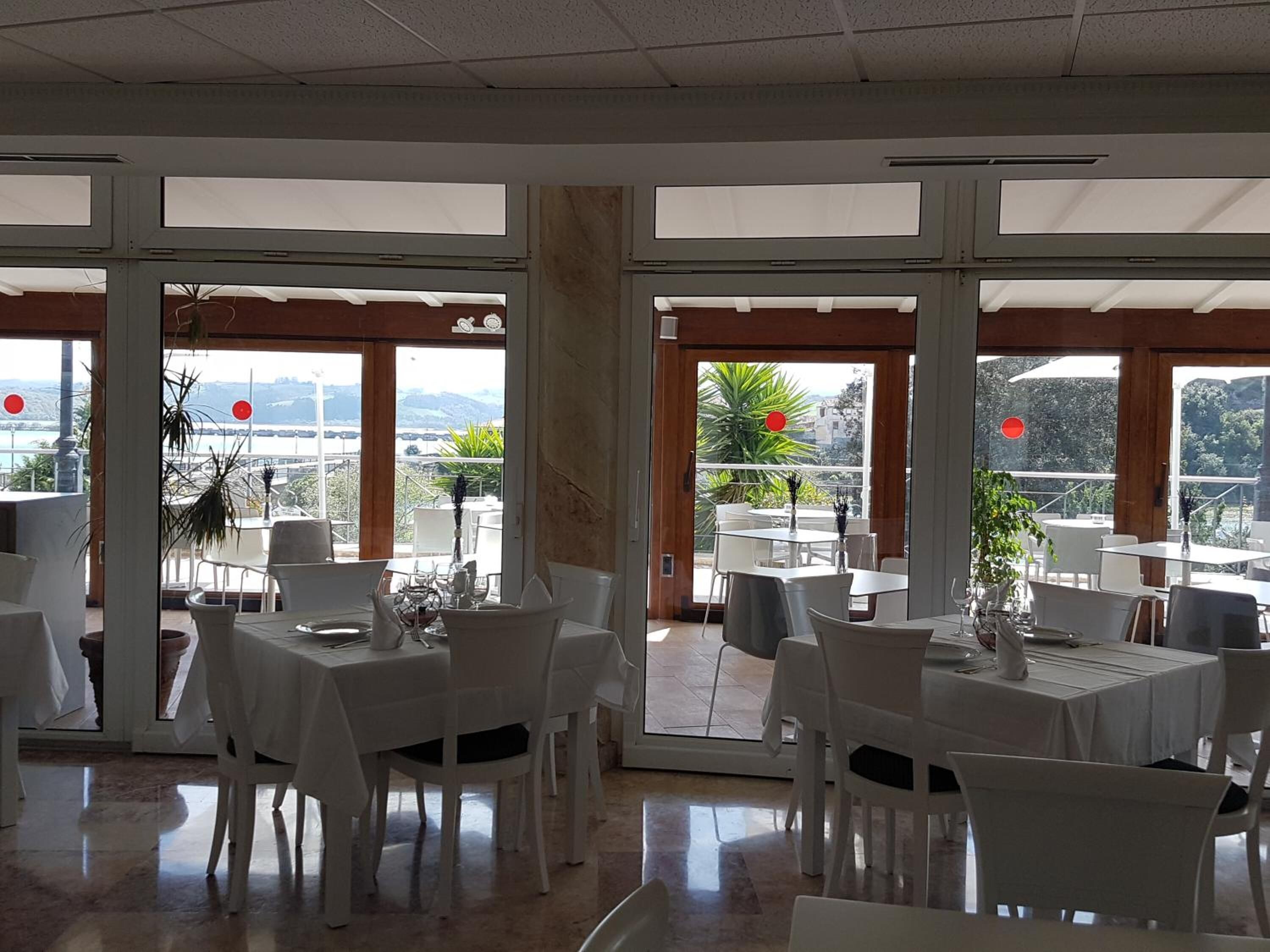 Un restaurante con varias mesas y sillas, ventanas amplias y vistas al exterior.