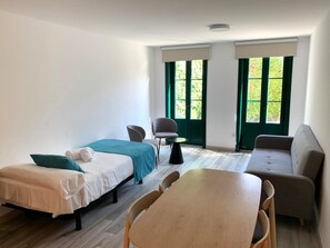 A room with two beds, a sofa, a table, and two windows with green curtains. - Pombal Rooms (Santiago de Compostela)