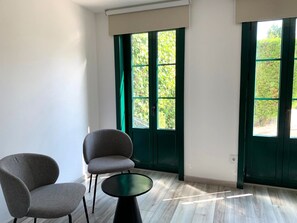 Una habitación con dos sillas y una mesita pequeña, ventanas amplias con marcos verdes y vistas al exterior con mucha vegetación.