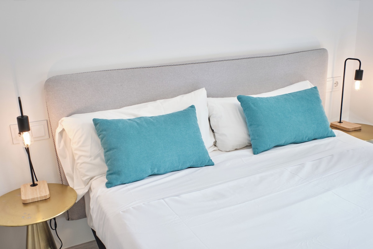 A neatly made bed with white and teal pillows, a grey headboard, and bedside lamps on wooden nightstands.