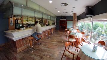 Zona de bar con taburetes, una barra y una persona trabajando detrás. Hay mesas y sillas en el comedor con vistas al exterior.